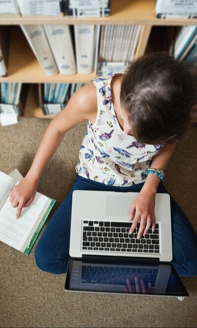 High angle view of a female student sitting against bookshelf with laptop on the library floor-1